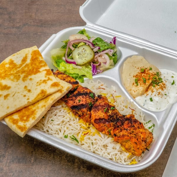 Grilled chicken skewers with rice, pita bread, salad, hummus, and tzatziki in a takeout container.