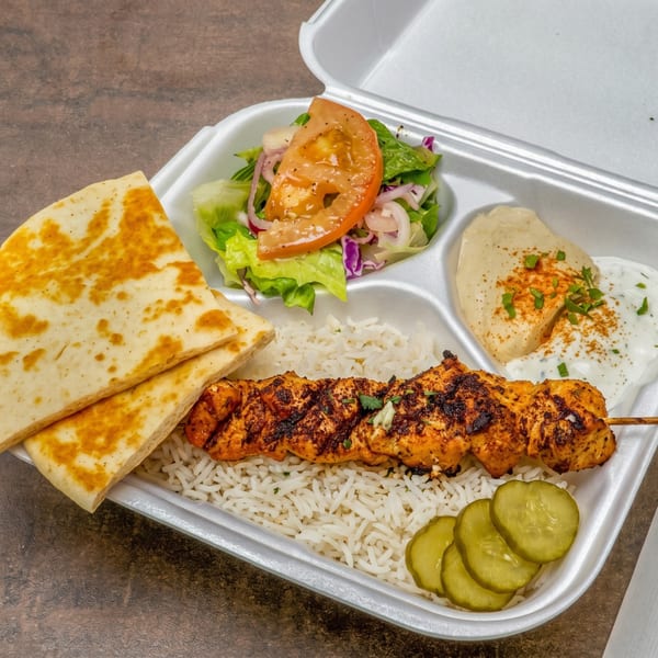 Grilled chicken skewer, rice, salad, hummus, tzatziki, pickles, and pita bread in a takeout container.