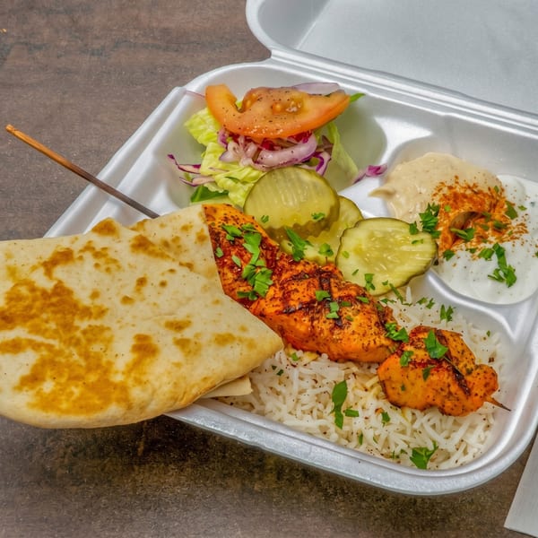 Chicken shish tawook with rice, pita bread, salad, pickles, hummus, and garlic sauce in a takeout container.